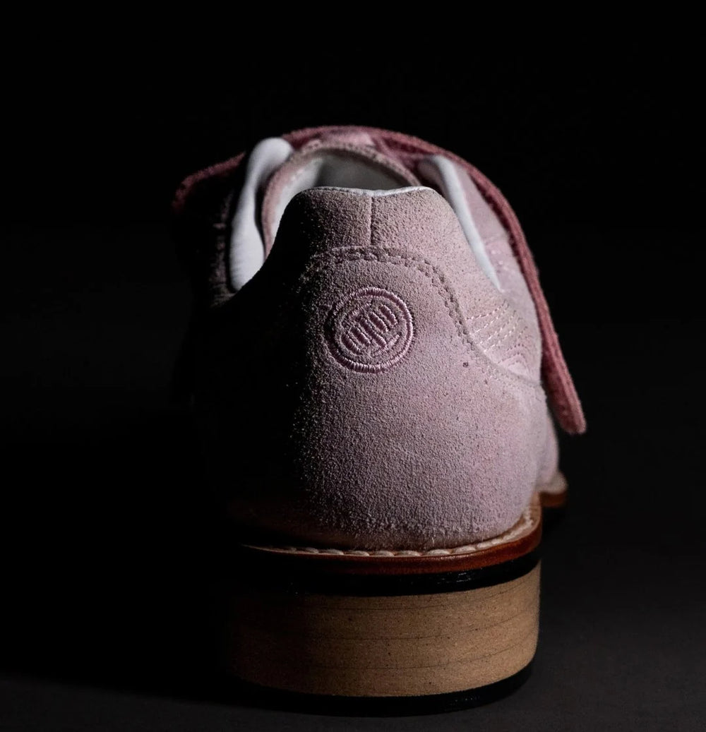 A close-up rear view of a dusty pink suede shoe with a wooden block heel, set against a solid black background.